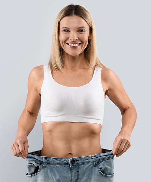 A smiling woman in a white sports bra holding out the waistband of oversized jeans, showcasing weight loss.