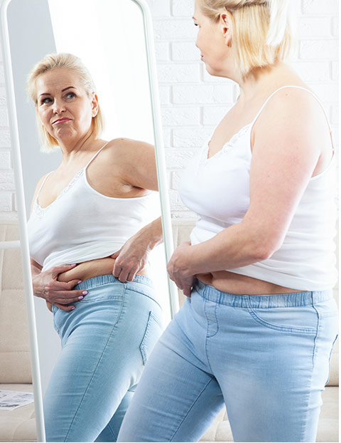 A middle-aged woman wearing a white tank top and jeans looks at herself in the
                mirror, pinching her waist and appearing thoughtful.