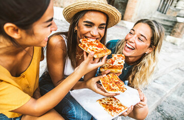 Three young women sitting outdoors and laughing while sharing slices of pizza.