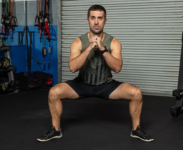 Coach Brian performing a sumo squat exercise in a gym environment, wearing a
                tank top and shorts.