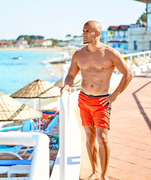 A fit, bald man in red swim trunks standing on a boardwalk by the beach, gazing into the distance.