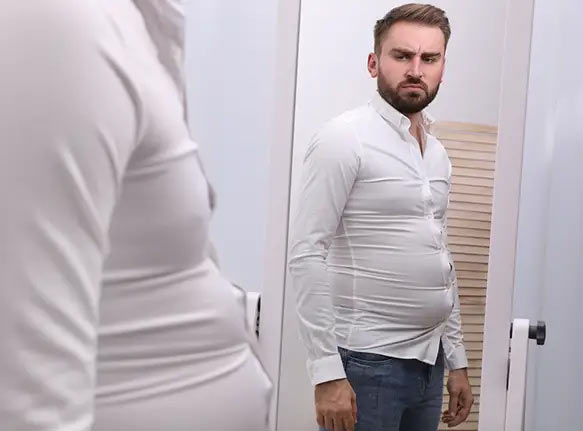 A man in a tight white shirt looking at his reflection in the mirror with a dissatisfied expression.