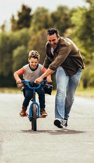 A fun and active day of a man helping a young child ride a bicycle on an outdoor path, with greenery in the
                 background.