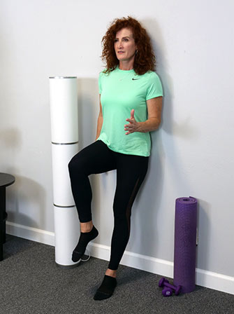 Tonya Fines performing an  exercise against a wall. She is standing on one leg, with foam rollers and a purple yoga mat and dumbbell nearby