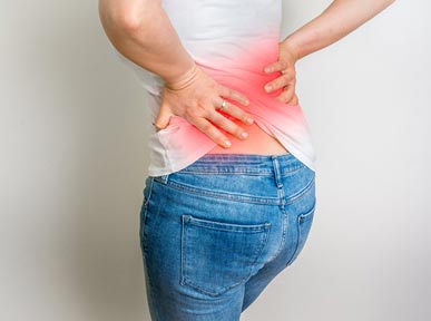 A woman in a white shirt and jeans holding their lower back, which is highlighted with a red
            overlay, indicating pain or discomfort in that area.